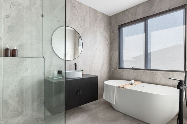 Contemporary bathroom with a freestanding white bathtub beside a large dark-framed window, a glass-enclosed shower, round wall mirror, floating dark vanity, and pale grey stone-look wall and floor finishes. The space has a clean, refined, spa-like appearance with abundant natural light.