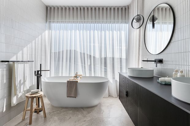 Bright contemporary bathroom featuring a freestanding white bathtub, double vessel basins on a dark fluted vanity, large oval mirror, wall-mounted black tapware, and white vertically ribbed wall tiles. Sheer curtains provide soft daylight, while pale stone-look flooring and restrained accessories create a refined, airy atmosphere.