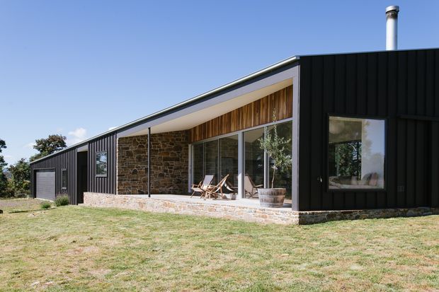 Single-storey contemporary rural house with a long, low-pitched roof and black vertical metal-clad wings. A recessed covered outdoor living area features exposed natural stone walls, warm timber lining, full-height glazing and a stone-paved terrace. The home sits in an open grassy setting under a clear sky.