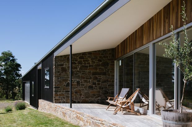Exterior view of a low-slung contemporary house with a broad covered terrace. The facade combines dark vertical metal cladding, natural irregular stone masonry, warm vertical timber wall cladding, and extensive aluminium-framed glazing. Timber lounge chairs sit on the paved terrace, with landscaped planting around the building and a clear blue sky.