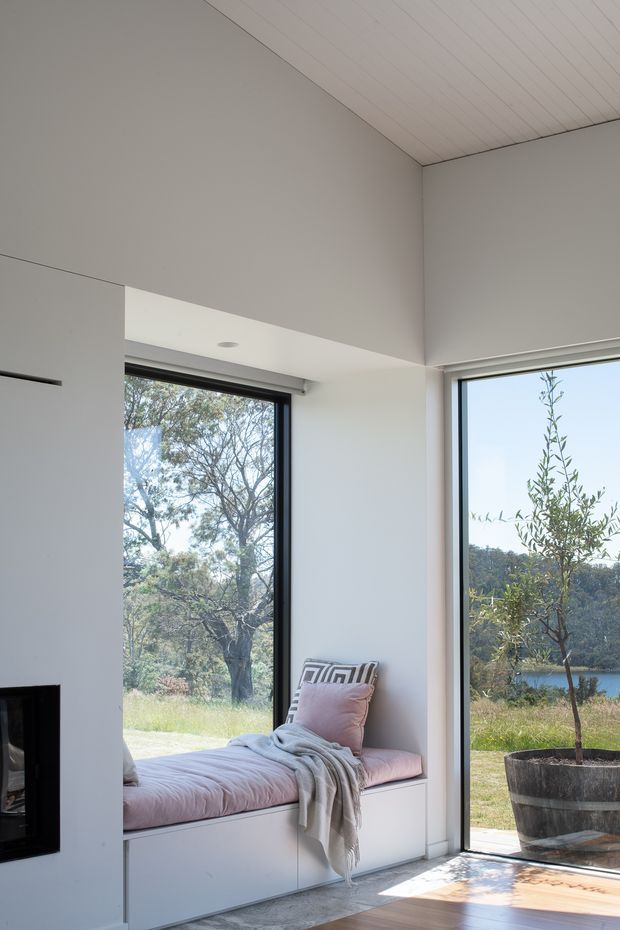 A bright minimalist interior corner featuring a built-in window seat with white storage cabinetry, soft pink cushions, a patterned pillow and draped throw. Full-height black-framed glazing opens the room visually to a landscaped outdoor setting with a potted tree and water beyond. The room has white walls, a pale slatted ceiling, light timber flooring, and a partially visible black fireplace at left.