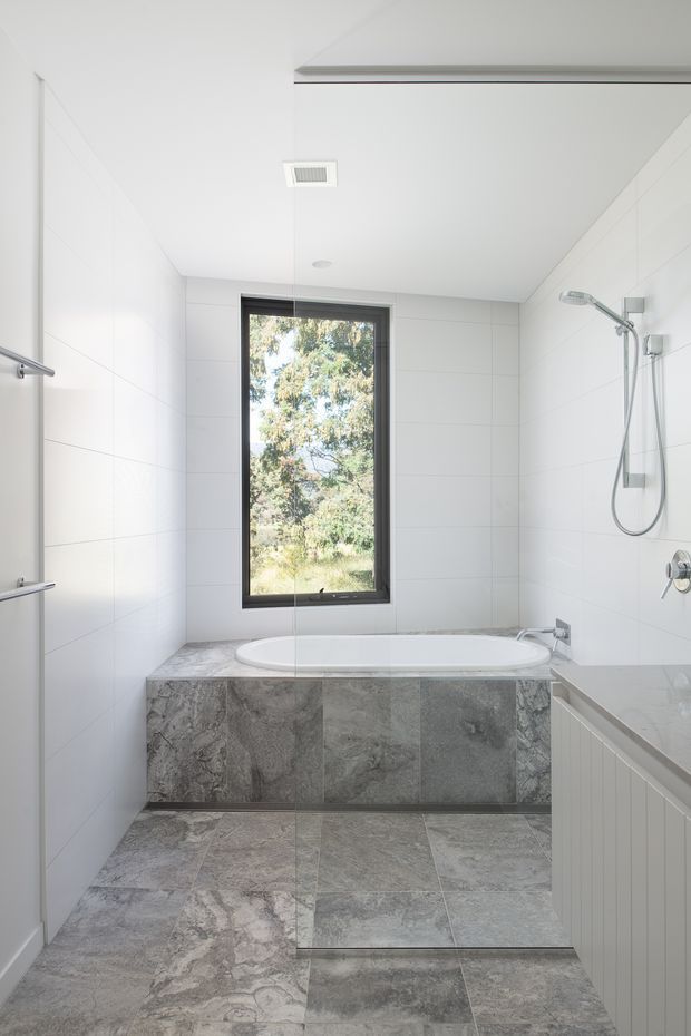 Bright, contemporary bathroom with a freestanding-style bathtub set into a grey stone-look tiled surround, matching large-format floor tiles, white wall tiles, a frameless shower area with chrome fixtures, and a dark-framed window overlooking trees. A partial vanity or built-in cabinet is visible in the foreground.