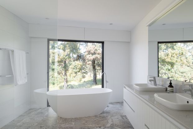 Bright contemporary bathroom with a freestanding white bathtub positioned in front of a large dark-framed window overlooking trees. A long white vanity with twin vessel basins, wall-mounted mirrored cabinetry, chrome fixtures, and white tiled walls fills the right side. The floor has a grey and beige natural stone-like finish, creating a clean, airy and tranquil atmosphere.