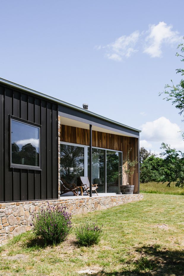 Exterior view of a compact contemporary single-storey building set in a landscaped rural setting. The facade combines dark vertical cladding, a low sloping roof, extensive glazing, warm timber lining beneath the covered terrace, and a rough natural-stone base. Outdoor chairs sit on the raised terrace, with lawn and planting in the foreground.