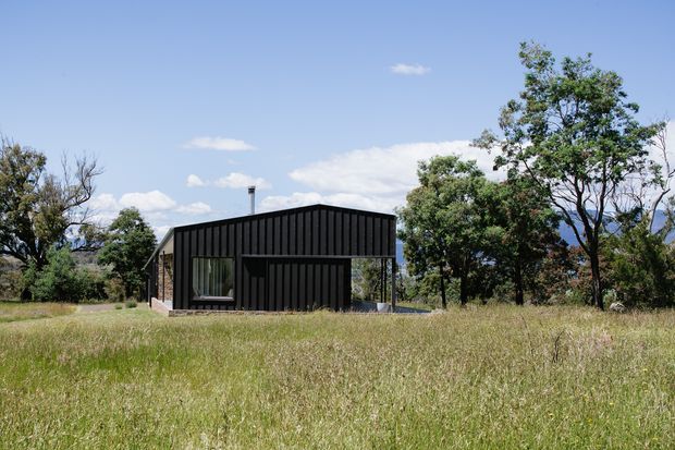 A compact single-storey black-clad house set within an open grassy rural landscape. The building has a simple contemporary rectangular form with a low pitched or skillion-like roof, vertical dark cladding, large glazed openings, and mature trees surrounding the site under a clear blue sky.