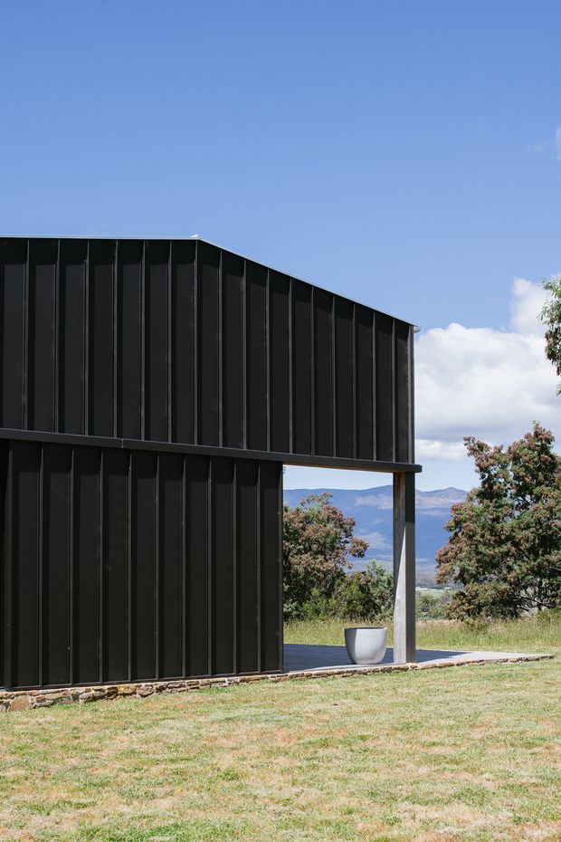 A minimalist contemporary two-storey building clad in dark vertical panels, photographed from the exterior. A large open ground-level bay frames a rural landscape with trees, distant mountains and blue sky. The building sits on a low rough-stone foundation, with a simple paved terrace and a large white planter visible beneath the overhang.