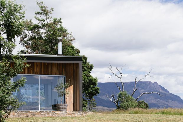 A compact modern cabin set in a natural landscape, featuring a dark flat roof, vertical timber cladding, broad floor-to-ceiling glazed sliding doors, and a cylindrical metal flue. The structure sits on a low stone base beside dry grass and trees, with dramatic mountain ranges visible beyond.