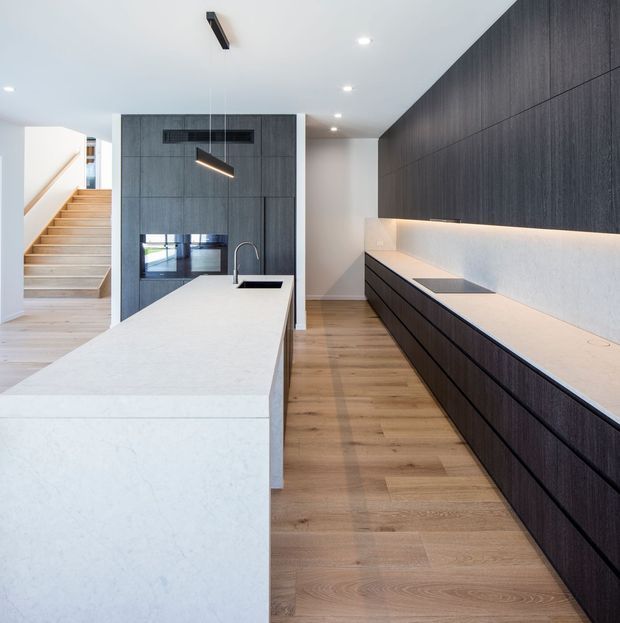 A contemporary galley-style kitchen with a large white stone island, dark vertically grained timber cabinetry, integrated under-cabinet lighting, and timber flooring. A staircase and adjoining circulation space are visible beyond the kitchen. The room has a clean, highly refined minimalist appearance with recessed ceiling lights and a modern linear pendant.
