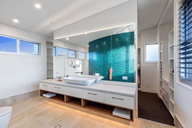 Bright contemporary bathroom with a long floating double vanity, white vessel basins, mirrored cabinetry, and a striking turquoise mosaic-tile feature wall. Large windows and white shelving provide abundant natural light, while pale timber-look flooring and warm under-vanity lighting add warmth. A doorway leads to a darker adjoining area.