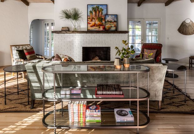 A bright, warmly styled living room centered on a white-painted brick fireplace with a dark timber mantel. The room features exposed dark ceiling beams, white walls, timber flooring, French doors and windows, patterned upholstered seating, a glass-and-metal console table, woven rug, framed artwork, and natural decorative elements. The overall look blends relaxed traditional architecture with eclectic, earthy furnishings.
