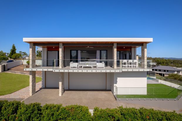 A contemporary two-storey residential house viewed from the front, featuring a flat roof with broad white fascia, exposed stone-clad structural columns, extensive upper-level glazing, a cantilevered balcony with clear glass balustrade, and an integrated garage at ground level. The home sits on a landscaped suburban site with lawn and neighboring houses visible in the background.