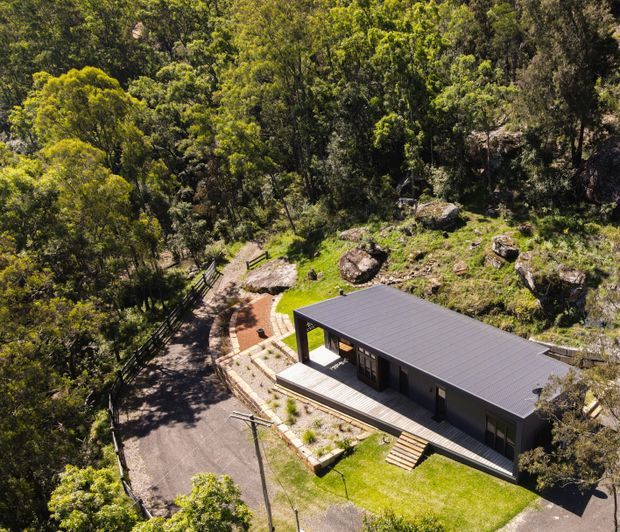 Aerial view of a contemporary single-storey dark-clad building set into a densely wooded, rocky sloping site. The building has a long low rectangular form, a broad flat roof, dark exterior walls, timber decking, paved paths, planted garden beds and a small manicured lawn.