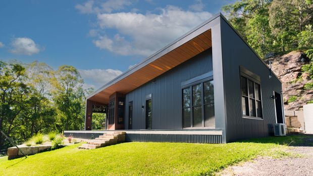 Exterior view of a compact contemporary single-storey house set on a sloping, wooded site. The building has a strong asymmetrical pitched roof, dark vertical metal cladding, large black-framed windows, and a warm timber-lined recessed soffit over the entry terrace. A small landscaped lawn and gravel driveway sit in front of the home.