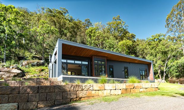 Exterior view of a contemporary single-storey house set among wooded landscaping. The building has dark vertical cladding, extensive glazing, a sharply sloping asymmetrical roof with a timber-lined soffit, and a sandstone block retaining wall in the foreground.
