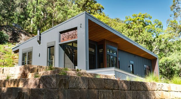 A modern single-storey house set into a wooded, sloping site. The compact building has dark grey vertical metal cladding, a broad asymmetrical roof with a deep timber-lined soffit, large windows and glazed doors, and substantial natural stone retaining walls in the foreground.