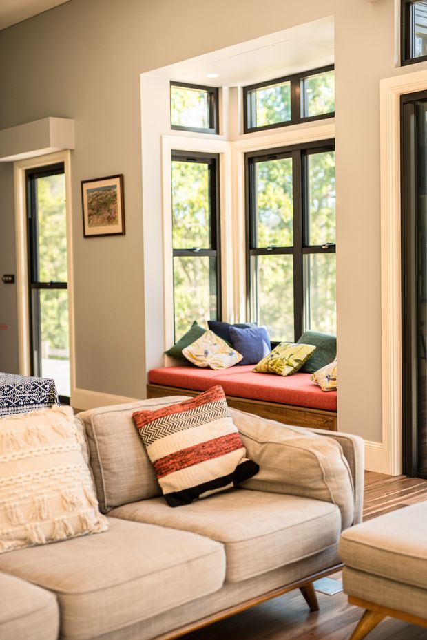 A warmly lit living room with a large upholstered sectional sofa and ottoman in the foreground, layered patterned cushions and throws, and a built-in window seat with a red cushion. The room features light neutral walls, dark timber flooring, black-framed multi-pane windows, and white architectural trim. A framed artwork and adjacent glazed openings are also visible.