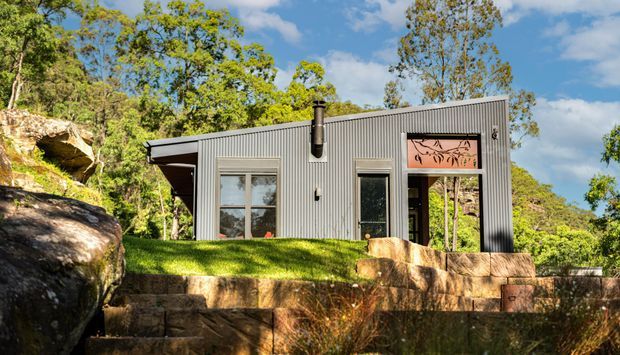 A compact contemporary pavilion or studio set into a leafy, rocky landscape. The building has a simple rectangular form, corrugated grey metal cladding, a low mono-pitched roof, large windows, and a glazed entrance with decorative artwork above it. Stone retaining walls and exposed rock surround the structure.
