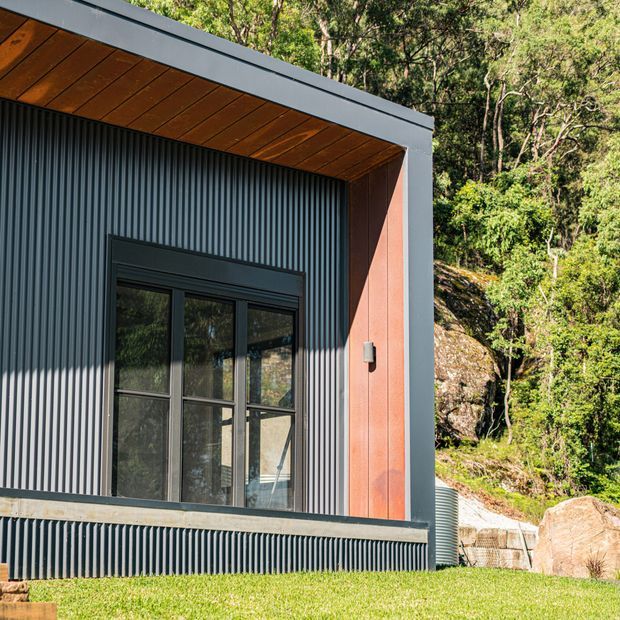 Close exterior view of a contemporary building set against a wooded, rocky landscape. The facade combines dark vertically corrugated metal cladding, a charcoal metal-framed glazed door or window, a warm timber-lined soffit, and a contrasting reddish vertical panel within a projecting rectangular roof and wall frame.