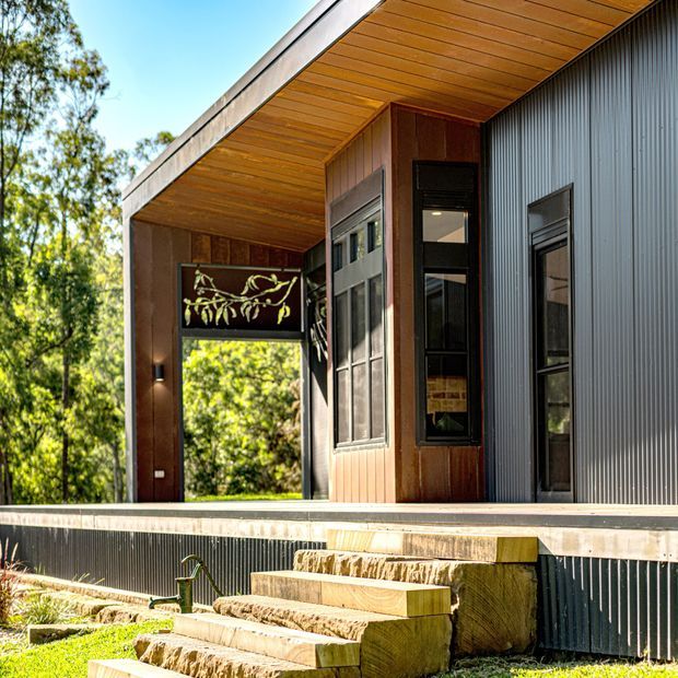 Exterior view of a contemporary single-storey building set among trees, featuring dark corrugated metal cladding, warm timber-lined soffits and timber wall panels, large dark-framed windows, and broad stone steps leading to a raised entry deck.