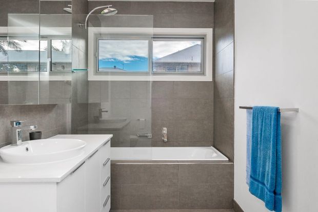 Contemporary bathroom with a white vanity and integrated basin, wall-mounted mirrored cabinetry, a tiled bathtub surround, and a white bathtub beneath a horizontal window. Large grey-brown tiles cover the walls and bath front, while a bright blue towel provides the main accent. A neighboring roof and outdoor scenery are visible through the window.