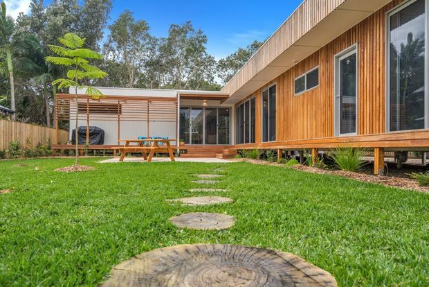Exterior view of a contemporary single-storey house set within a leafy landscaped garden. The building has warm vertical timber cladding, extensive glazing, a long low roofline, raised timber decking, and a timber pergola enclosing an outdoor dining area. Circular timber stepping stones lead across the lawn toward the house.