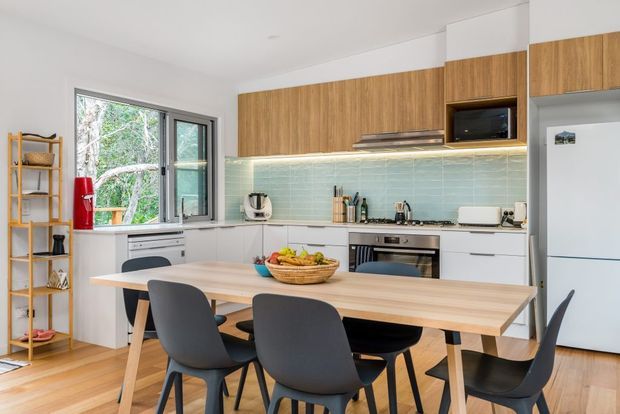 Bright contemporary open-plan kitchen and dining area with white lower cabinetry, warm timber overhead cabinets, a pale aqua tiled splashback, stainless-steel appliances, and a large light timber dining table surrounded by dark grey chairs. A window brings in greenery, while open shelving and a white refrigerator complete the space.