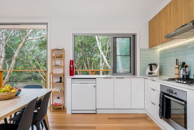 Bright contemporary kitchen and dining area with white cabinetry, timber upper cabinets, pale aqua tiled splashback, stainless-steel appliances, timber flooring, and large windows overlooking trees. A dining table and chairs occupy the foreground, while open shelving and integrated appliances line the kitchen.