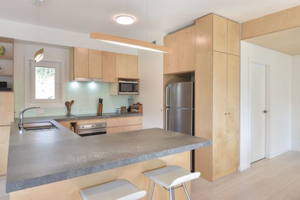 Bright contemporary kitchen with extensive light timber cabinetry, a large grey island benchtop with integrated seating, stainless-steel appliances, pale timber flooring, and white walls. A window provides natural light, while simple pendant and ceiling lighting create a clean, understated interior.