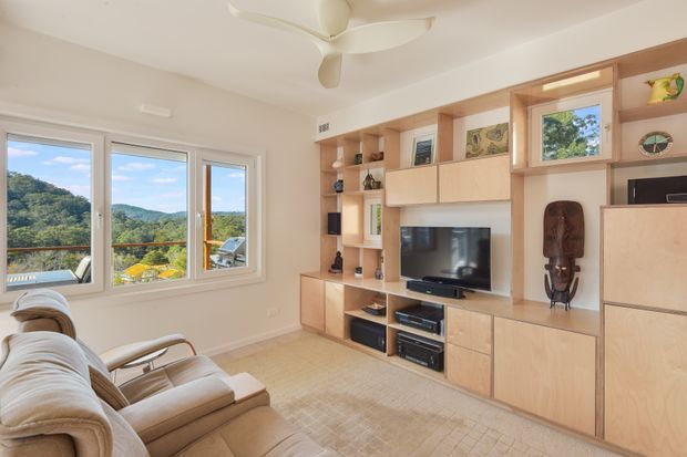 Bright living room with a large built-in timber entertainment and storage wall featuring open shelving, cabinetry, and a television. A beige sectional sofa and armchairs face the media unit, while wide windows provide expansive views of a green hillside and water beyond. The room has a light neutral palette, a ceiling fan, and integrated display niches.