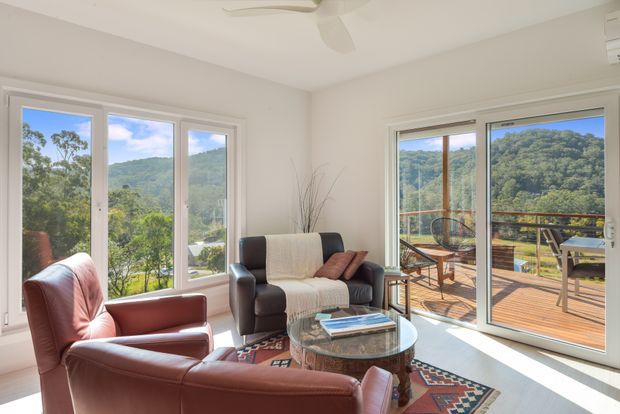 Bright living room with large white-framed windows and sliding glass doors opening onto a timber deck. The room contains two brown leather armchairs, a dark sofa with a throw, a round glass coffee table, and a patterned rug. Lush wooded hills are visible outside, creating a leafy outlook and strong indoor-outdoor connection.