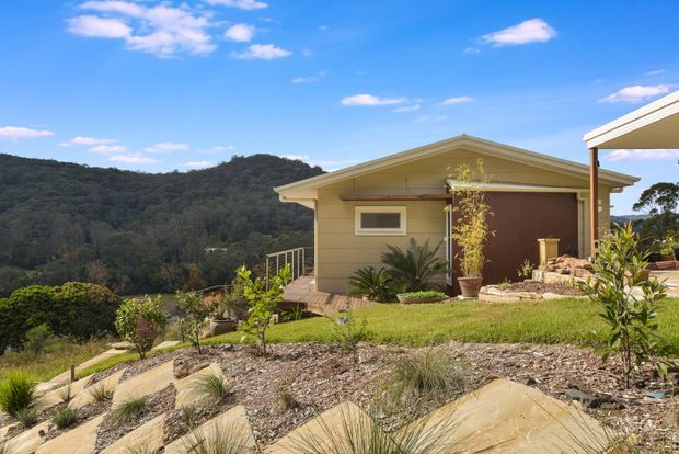 A compact single-storey house or pavilion set on a landscaped sloping site, viewed from outdoors. The building has a simple gable roof, pale horizontal exterior cladding, a dark brown entry door, and a small deck with metal railing. Native-style planting, landscaped garden beds, and stone-edged paths surround the structure, with wooded hills in the background under a bright blue sky.