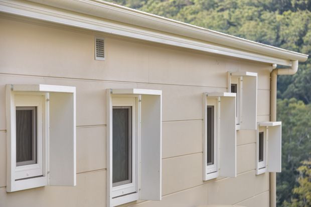 Close-up exterior view of a pale cream rendered or panelled building facade with a row of narrow windows, each fitted with projecting white external shutters or protective surrounds. The low-pitched roof has white eaves and gutters, and a wooded hillside is visible in the background. The image focuses on the building envelope and window treatment.