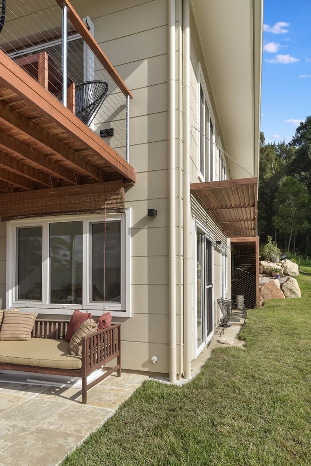 Exterior view of a multi-storey residential building with pale horizontal weatherboard-style cladding, white-framed glazing, timber balconies and decks with cable balustrades, and a covered outdoor seating area. The building sits beside a landscaped grassy slope with exposed rocks and trees beyond.