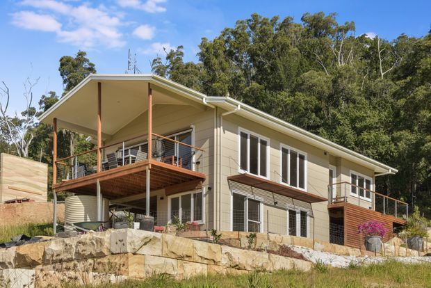 Exterior view of a contemporary elevated multi-level house set into a sloping, heavily wooded site. The home has pale horizontal cladding, a low-pitched roof with broad eaves, extensive glazing, slender timber-toned posts, and several timber decks with metal balustrades. A stone retaining wall and rainwater tank are visible below the house.