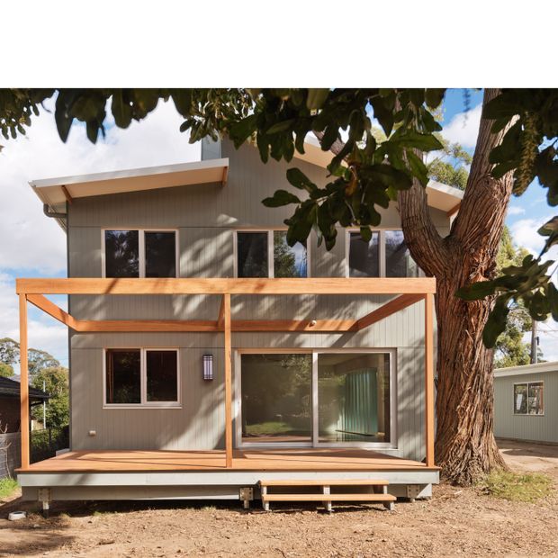 Exterior view of a compact two-storey contemporary dwelling set among mature trees. The facade combines vertical grey cladding with white trim, a low-slope asymmetrical roof, large windows and glazed sliding doors. A raised timber deck with simple timber-framed screening and central steps spans the front elevation.