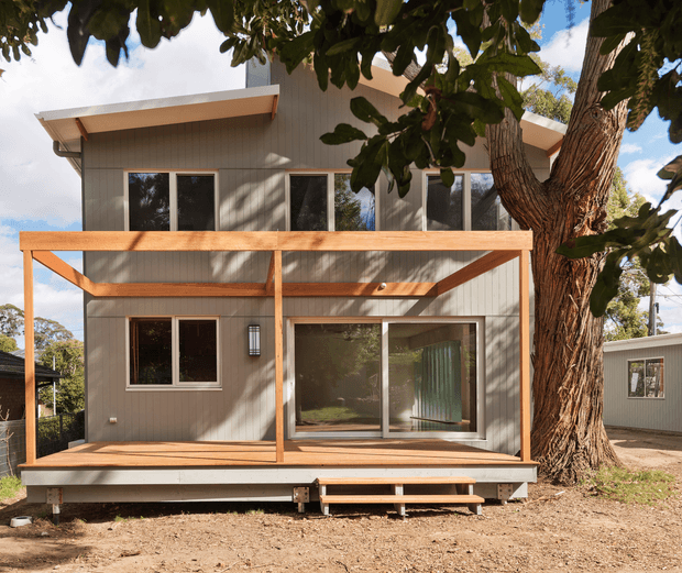 Exterior view of a compact two-storey contemporary dwelling set among mature trees. The facade combines grey vertical cladding with large dark-framed windows and sliding glazing, while a broad timber-framed covered deck and steps occupy the lower level. A low-pitched projecting roof and simple rectangular form create a clean, modern cabin-like appearance.
