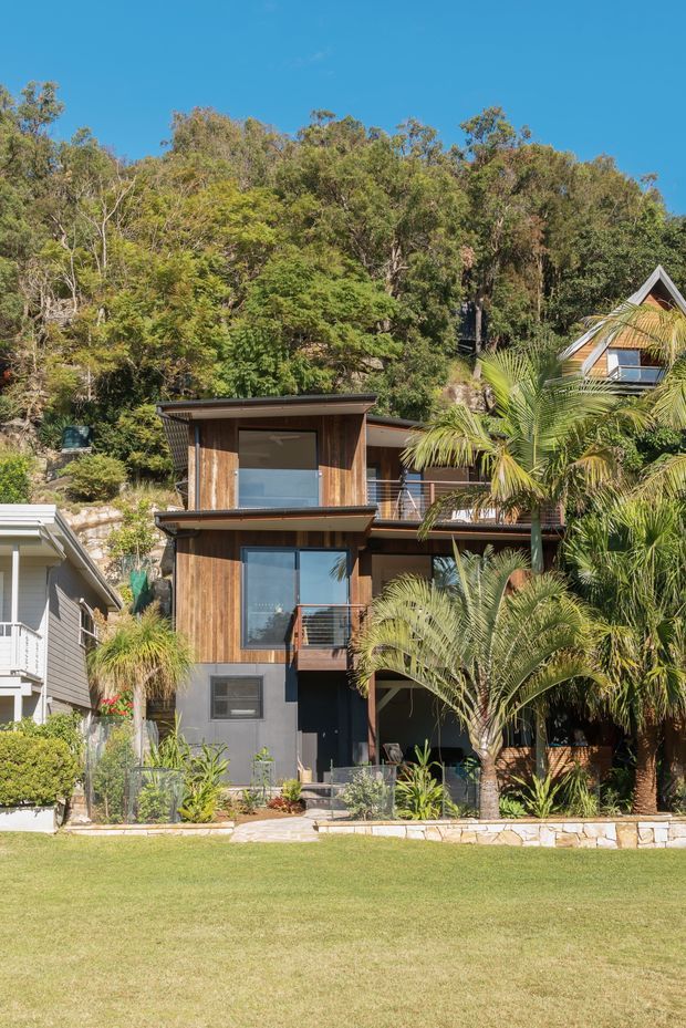 Exterior view of a compact multi-storey contemporary home set into a lush, wooded hillside. The facade combines vertical timber cladding, dark painted lower walls, expansive glazing, slim metal balcony railings, and shallow projecting roof forms. Tropical palms and landscaped planting occupy the foreground, with neighbouring homes partially visible at either side under a clear blue sky.