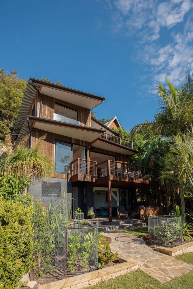Exterior view of a multi-storey residential house set within lush tropical landscaping. The home features dark timber cladding, broad projecting roof eaves, multiple balconies and decks with horizontal metal balustrades, large glazed openings, and a stone-paved garden path. A bright blue sky and dense palms and subtropical planting surround the elevated-looking structure.
