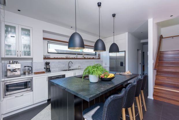 Bright contemporary kitchen with white full-height cabinetry, integrated appliances, a large dark stone island with bar seating, and three matte dark pendant lights. A timber-framed horizontal window sits above the marble-look splashback and sink, while a timber staircase is visible at right. The space has a clean, refined look with contrasting dark flooring and joinery.