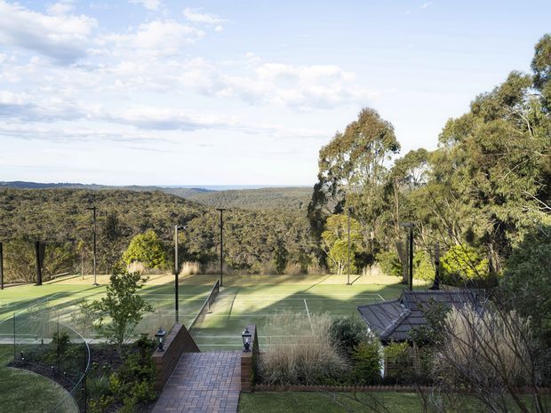 Elevated outdoor view over a landscaped property with multiple fenced tennis courts, a brick-paved walkway, garden planting, mature eucalyptus trees, and a wooded rural landscape extending to the horizon. The scene is bright and open, with the recreational courts and surrounding grounds as the main focus.