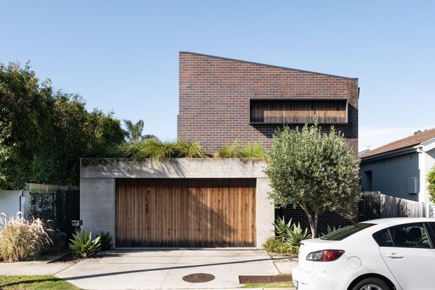 Street-facing contemporary residence with a strong geometric form, red-brown brick upper volume, concrete garage structure, and a projecting dark-framed window with vertical timber screening. The lower garage door also uses vertical timber battens, while drought-tolerant planting softens the paved frontage. A neighboring house and parked car are visible but secondary.