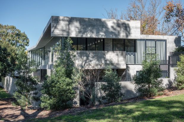 Exterior view of a contemporary multi-level residence with a strong horizontal composition, flat roof, extensive dark-framed glazing, and pale textured concrete or rendered facade surfaces. Mature landscaping partially screens the building, while projecting upper volumes and recessed balconies create depth and shadow.