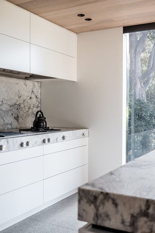 A bright, refined contemporary kitchen with streamlined white handleless cabinetry, a marble splashback and matching stone benchtops. A stainless-steel range cooker and black kettle sit beneath a concealed rangehood, while a timber-lined ceiling and full-height glazing bring warmth and views of trees into the space. The foreground shows the substantial edge of a marble island.