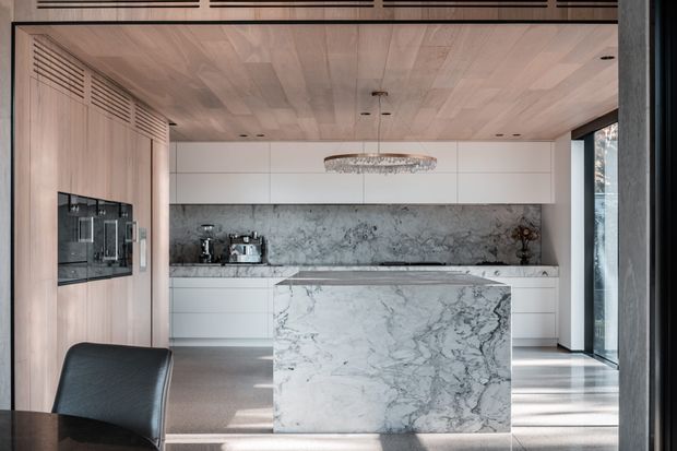 Contemporary open-plan kitchen with a large marble-clad island, white handleless cabinetry, a dark veined stone splashback, pale timber-lined ceiling and wall, integrated appliances, and full-height glazing. A sculptural linear pendant and dark dining chair add contrast, while strong natural light creates a refined, high-end atmosphere.