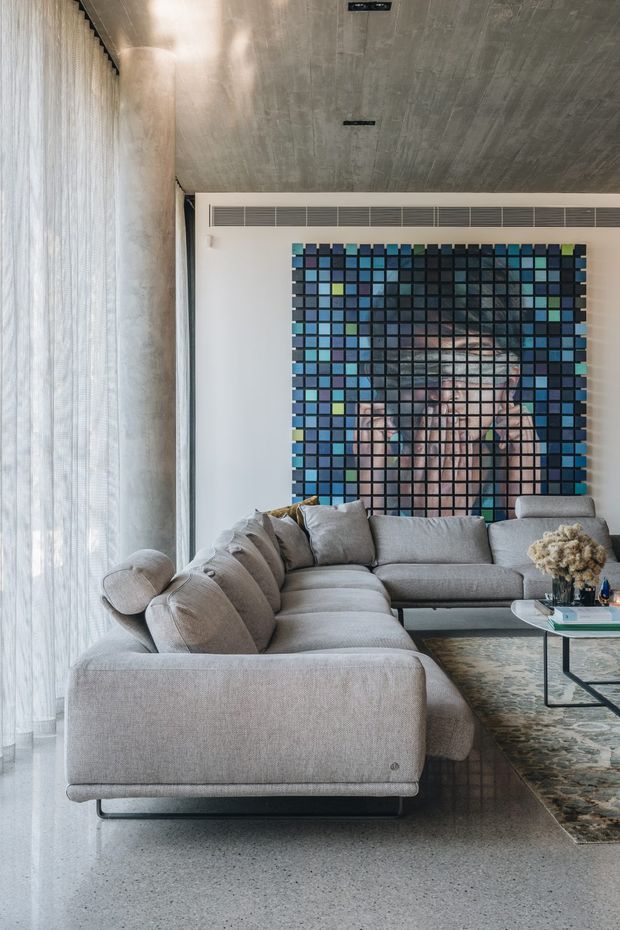 A refined contemporary living room with a large pale grey sectional sofa, glass coffee table, patterned rug, sheer floor-to-ceiling curtains, and a striking blue and black grid-like artwork or feature installation on the rear wall. The ceiling has a raw concrete finish with recessed linear lighting, while the polished light-toned floor and restrained furnishings create a calm, gallery-like interior.