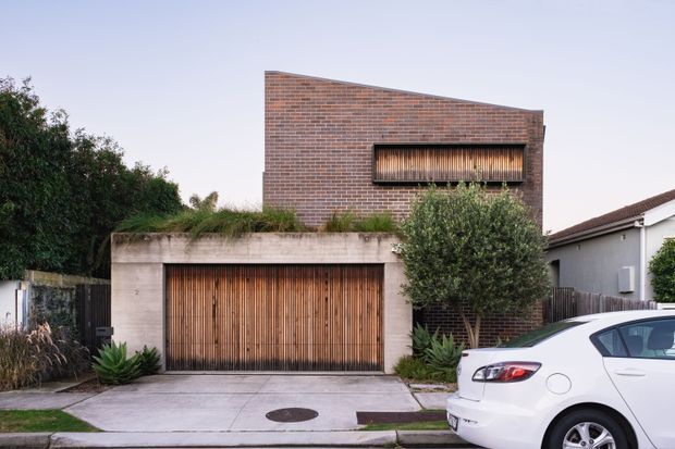 Street-facing residential facade with a strong geometric composition: a lower concrete garage volume with a vertically slatted timber garage door, topped by a planted roof terrace, and an upper red brick volume with a shallow sloping roofline and recessed horizontal window fitted with vertical timber screening. Mature planting softens the frontage, while a neighbouring house and parked car appear at the edges.
