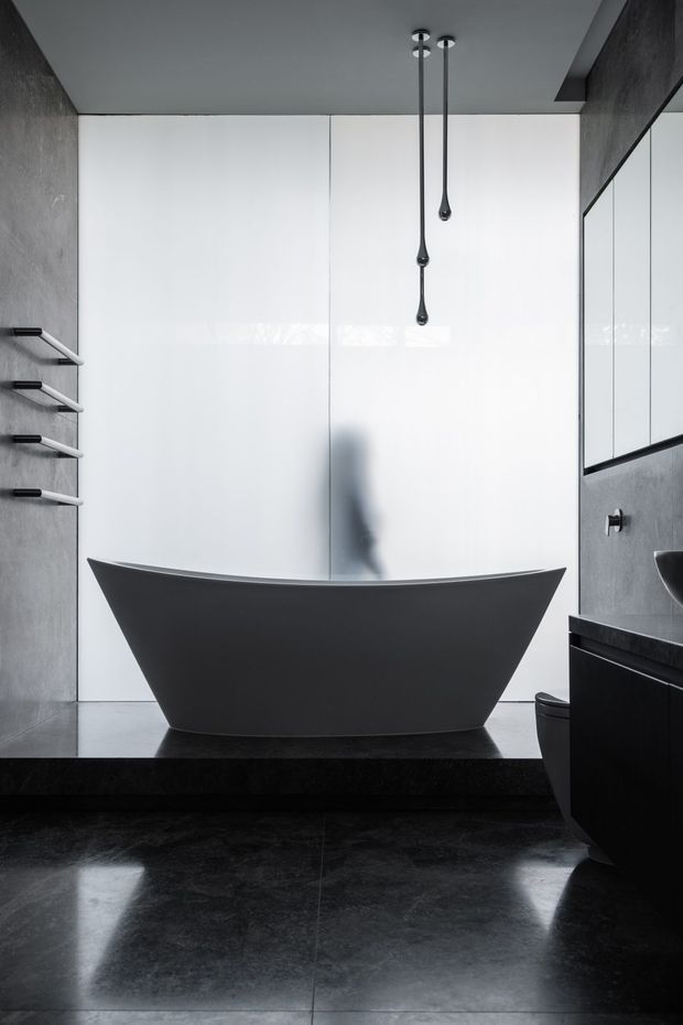 A dramatic contemporary bathroom dominated by a sculptural matte-black freestanding bathtub set against a large white wall or frosted-glass backdrop. Dark stone-look flooring, charcoal textured walls, black built-in cabinetry, a wall mirror, minimalist black tapware, and slender pendant lights create a monochromatic, high-contrast interior. A shower area and toilet are partially visible.