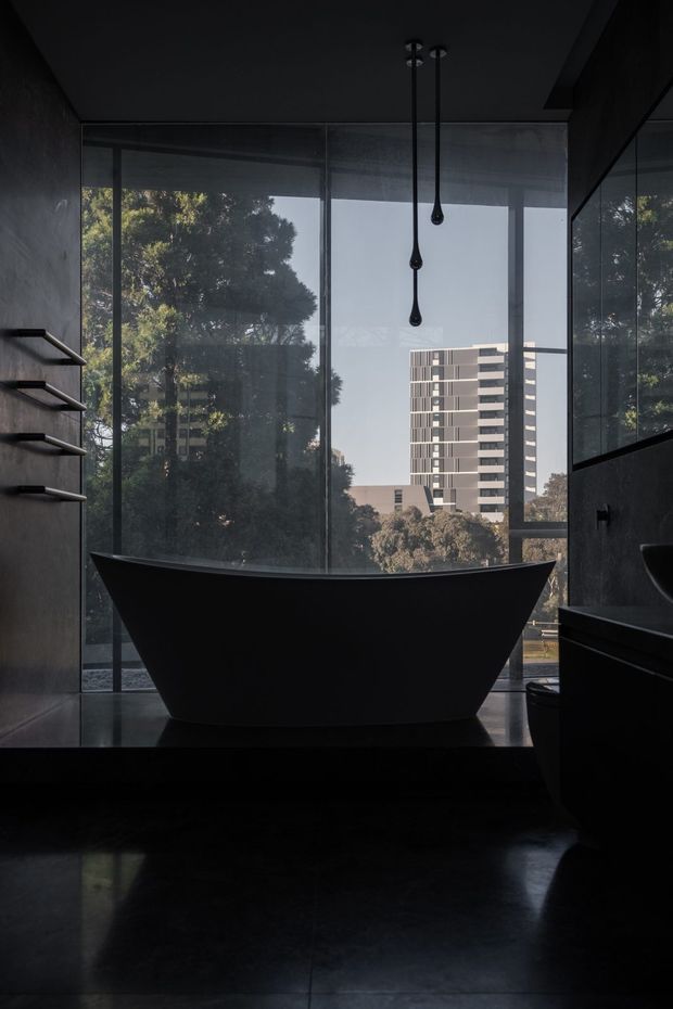 A dramatic, dark contemporary bathroom featuring a sculptural freestanding matte-black bathtub positioned in front of full-height glazing. The room includes dark wall and floor finishes, minimalist black tapware and shower fixtures, floating open shelving, and a dark vanity. Reflections in the glazing and mirror-like surfaces extend the sense of space, while trees and a modern apartment building are visible outside.