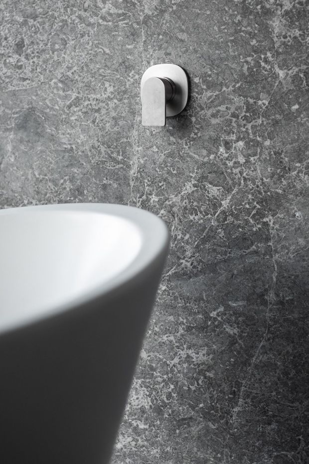 Close-up monochrome bathroom detail showing a freestanding white bathtub in the foreground, a minimalist wall-mounted bath control, and a dark grey veined stone wall finish.