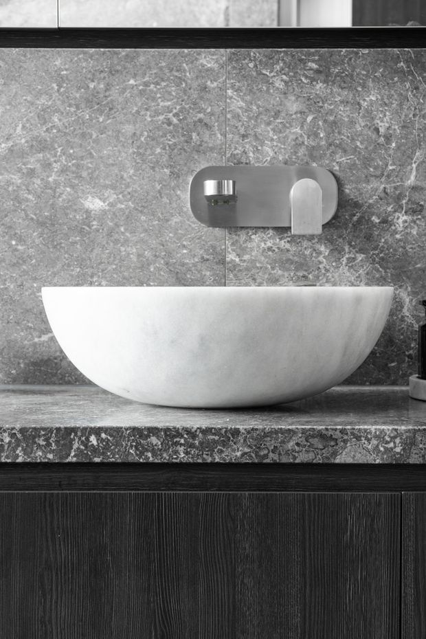 Close-up monochrome photograph of a contemporary bathroom vanity. A white oval vessel basin sits on a grey veined stone benchtop and backsplash, with a wall-mounted brushed metal tap above. Dark vertically textured timber cabinetry forms the vanity base, with dark trim and a partially visible mirror or upper surface.